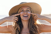Beautiful smiling woman applying sunscreen on her face while looking at camera at the beach.