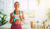 Close up image of beautiful happy excited young sporty slim woman carrying jumping rope on shoulders after home workout and looks on camera. Sport and recreation concept at home. Strong and power
