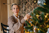 Portrait of elegant middle-aged woman hangs up ball on branch of xmas tree in cozy living room, looking at camera. Happy female house decoration