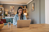 A young cheerful woman sitting at the table in the living room at home and using a laptop.