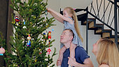 Young family decorates a christmas tree in their home, with the daughter perched on her father's shoulders to hang a christmas bauble on a high branch. New Year's Eve with family