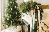 Stylish festive decorated fireplace mantel with fir branch, bells, ribbon on background of christmas tree. Festive christmas winter decor in modern scandinavian room. Merry Christmas!