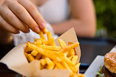 Closeup of young womans hand grabbing a crispy golden fries. Enjoying potato chips with a juicy burger in fast food restaurant on summer day. Selective focus