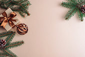 Christmas flat lay with green pine branches, bronze gift box with ribbon and ornaments arranged on a beige background. Top view, copy space